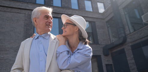 A woman in a hat and glasses hugs an elderly man in a white jacket from behind. Romantic relationships of mature people. 