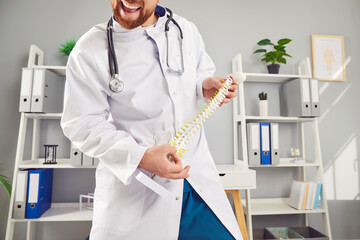 Funny young man doctor orthopedist standing in medical office. Male chiropractor or osteopath holding spine of human skeleton anatomical model in his hand. Health care and medicine concept.