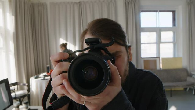 Handheld POV shot of young Caucasian male photographer using professional camera with big lens and flash while taking pictures in spacious sunlit studio working with colleague or assistant