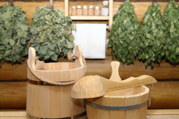 Traditional accessories for sauna and bath treatments  stand on wooden bench in sauna interior on the background of log wall with dry brooms and shelf with set of aroma oil bottles and white towel.