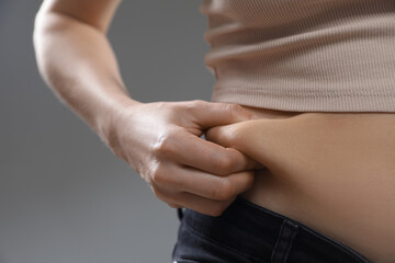 Woman touching belly fat on grey background, closeup. Overweight problem