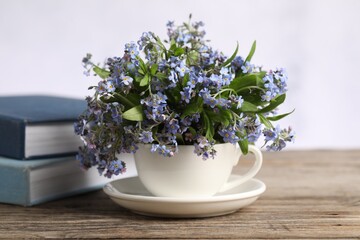 Beautiful forget-me-not flowers in cup, saucer and books on wooden table against light background,...