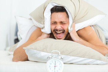 man covering ears with pillow to hide alarmclock noise