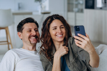 Joyful couple taking selfie together with smartphone, in bright and airy modern living space. Man makes funny mustaches out of woman's hair. 