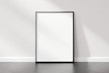 Blank white vertical poster frame standing against a white wall in a room with wooden flooring,...