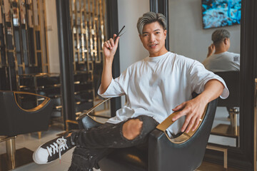 Happy Asian professional Hairdresser or hairstylist man sitting confidence with smile and holding hairdressing equipment while looking at camera in beauty salon and barber shop