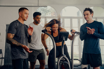 Multiracial young adults in sportswear applauding for friend cycling on exercise bike at the gym