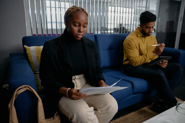 Two multiracial young business people in businesswear using digital tablet and reading on sofa in office
