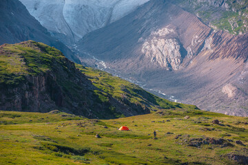 Vivid orange tent in sunlit vast alpine valley among green hills and rocks with view to big glacier...