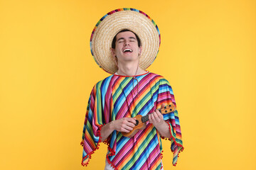 Young man in Mexican sombrero hat and poncho playing ukulele on yellow background