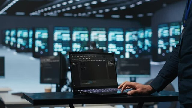 IT support employee writing code on laptop in high tech facility with server rows providing computing resources for different workloads. System administrator uses notebook to oversee supercomputers