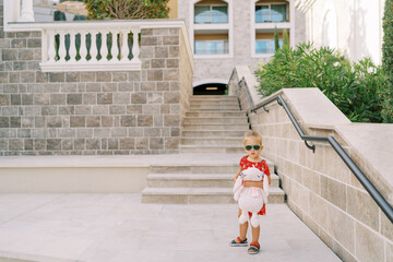 Little girl with a pink toy hare stands near the stairs of an apartment building in the garden