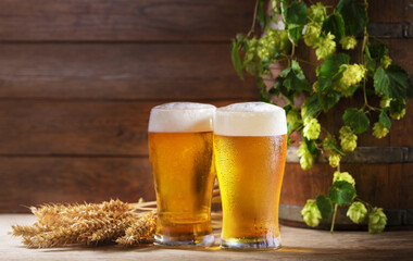 Glasses of beer, wooden barrel, wheat ears and green hops on wooden background