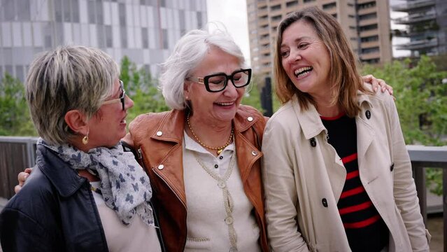 Group of three mature caucasian women enjoying embracing walk together laughing outdoor. Senior female friends having fun hugging strolling happy in the urban city street. Elderly people enjoying