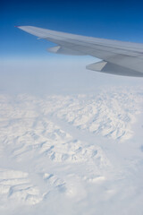 Obraz premium View from a flying airplane of snow-capped mountains under the wing of the airplane. Winter aerial landscape. Travel and aviation tourism in the Arctic. Flight on a passenger airliner. Polar region.