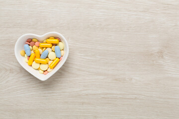 Medicines in heart-shaped bowl on wooden background, top view