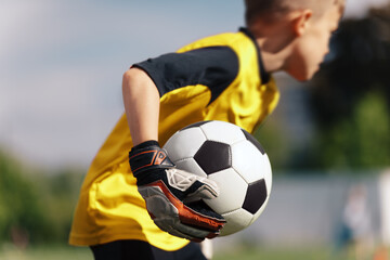 Young boy as a soccer goalie holding the ball in one hand ready to start a game. Football goalkeeper in jersey shirt and sports gloves throwing ball and playing a football match