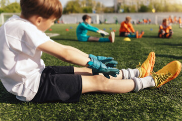 Young Boys as Football Goalkeepers Sitting on Soccer Pitch and Warming Up Before the Training Unit. Happy Kids in Stretching at Football Field. Kids Wearing Goalkeepers Uniforms and Soccer Gloves