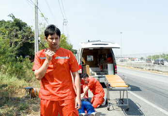 Asian rescue staff in orange uniform use walkie talkie to coordinate the nearest hospital, stand in front of an ambulance with open door and colleague working behind. © chadchai