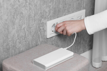 Woman plugging power bank into socket on grey wall indoors, closeup