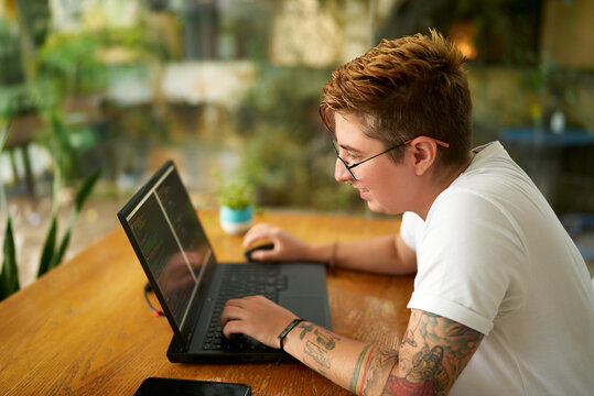 Transgender software developer programs at wooden desk. Focused on coding for mobile app, progressive workplace. Note tattoos, casual work attire. Green office signifies eco-friendly company culture.
