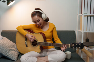 Young woman wears music headphones playing a musical instrument while practicing acoustic guitar...