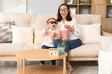 Beautiful mother and her cute little daughter in 3D cinema glasses with popcorn watching movie at home