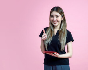 Young asian woman stand with a thumbs up, smile while holding tablet computer. Portrait on pink background with studio light. Isolated