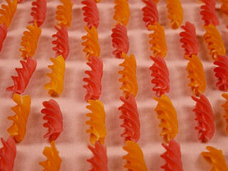 Italian uncooked orange and red pasta fusilli are laid out in rows on light pink fabric, natural...
