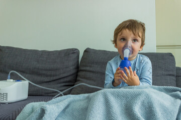 The boy makes inhalations with nebulizer at home, he's got a cold and a blanket.