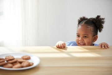 African child girl hiding and looking chocolate cookies or biscuits on dish from under the table