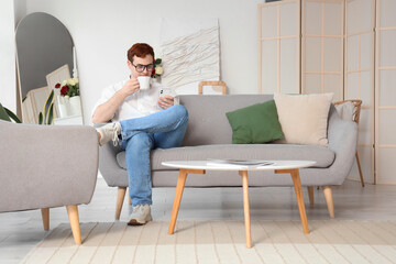 Obraz premium Young man in eyeglasses with cup of tea and mobile phone sitting on grey sofa at home