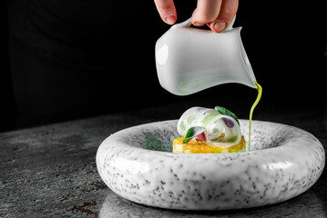 A chef’s hand pouring green sauce on an artistic gourmet dish in a speckled white plate against a dark background.