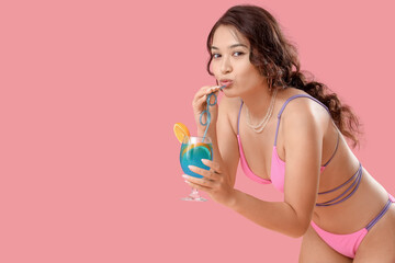 Sexy young woman in swimsuit and with Blue Lagoon cocktail on pink background
