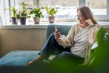 Caucasian attractive mature middle aged woman sitting on the couch at home using smartphone