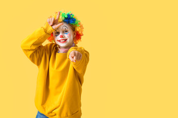Funny little girl in clown wig showing loser gesture on yellow background. April Fools' Day celebration