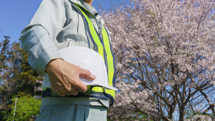 桜の花とヘルメットを持つ作業服姿の男性