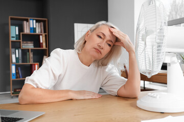 Mature businesswoman with electric fan suffering from hot flashes at desk in office. Menopause...