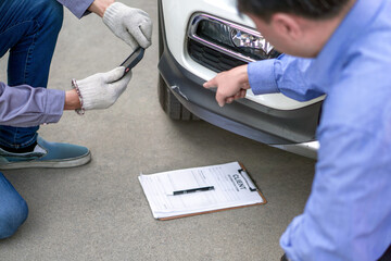 Two individual inspect damage on a vehicle. Car’s owner pointing at a dent on the bumper of the car. An insurance staff taking picture with smartphone, conducting an assessment related to the damage.