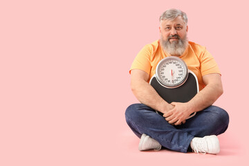 Overweight happy mature man with scales sitting on pink background. Weight loss concept