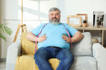 Overweight happy mature man with measuring tape sitting on sofa at home. Weight loss concept