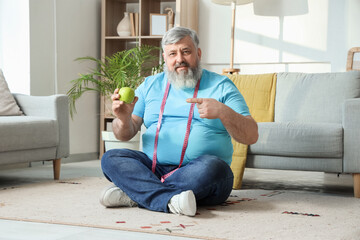 Overweight happy mature man with measuring tape pointing at apple in room. Weight loss concept
