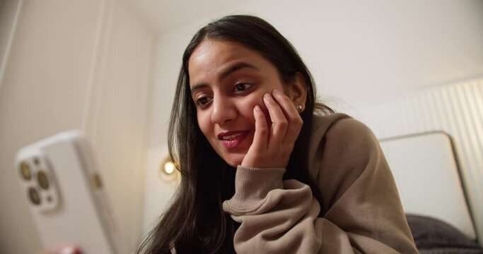 A confident brunette girl in a brown sweater communicates with her friends online using a video call on a white smartphone while lying on the bed in a modern apartment