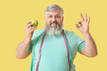 Overweight happy mature man with measuring tape showing OK gesture on yellow background. Weight loss concept