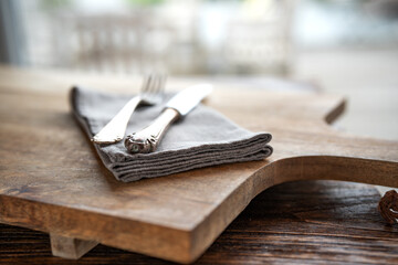 Simple rustic table setting on wooden cutting board. Old silver cutlery with linen napkin and black ceramic dishes.