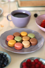 Plate of pastel macarons, cookies and chocolate, cup of tea of coffee, glass of bubble water, various berries, books and accessories on the table. Selective focus, pastel colors.