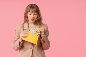 Surprised young woman holding yellow wallet and money on pink background