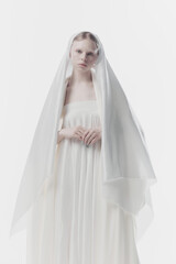 Young woman shrouded in gossamer white veil, creating ghostly yet angelic appearance. against white...