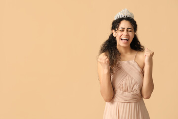 Emotional young African-American woman in stylish beige prom dress and tiara on brown background © Pixel-Shot