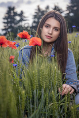 Portrait of beautiful long haired brunette tanned girl dressed in jeans sitting in cornfield with red poppies. Vertically. 